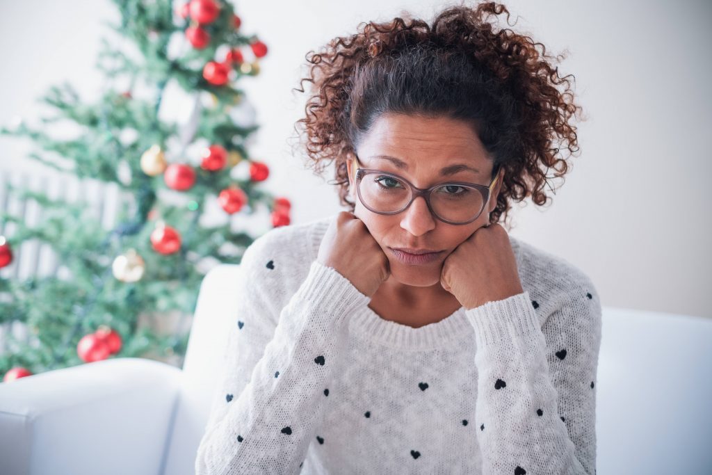 a young African American woman in a white sweater, wearing glasses, looking sad, disappointed, stressed and tired with her head resting on her hands and a holiday Christmas tree and decorations in the background