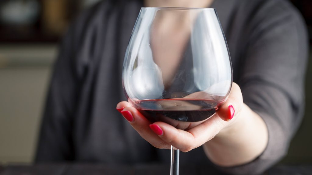 a Caucasian woman in a grey sweater sitting at a table, wearing red fingernail polish and holding a glass of red wine