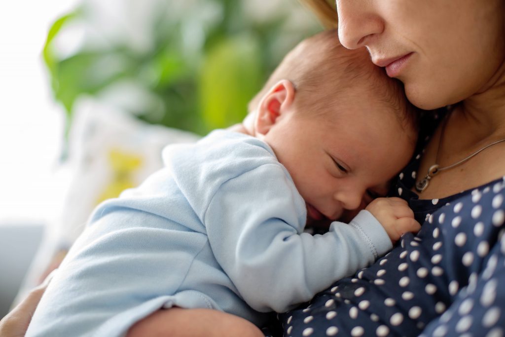 a young Caucasian woman, perhaps a mother, in a polka dot blouse, looking calm, serene, or maybe sad and concerned, holding or cuddling an infant, baby in a blue onesie near her chest