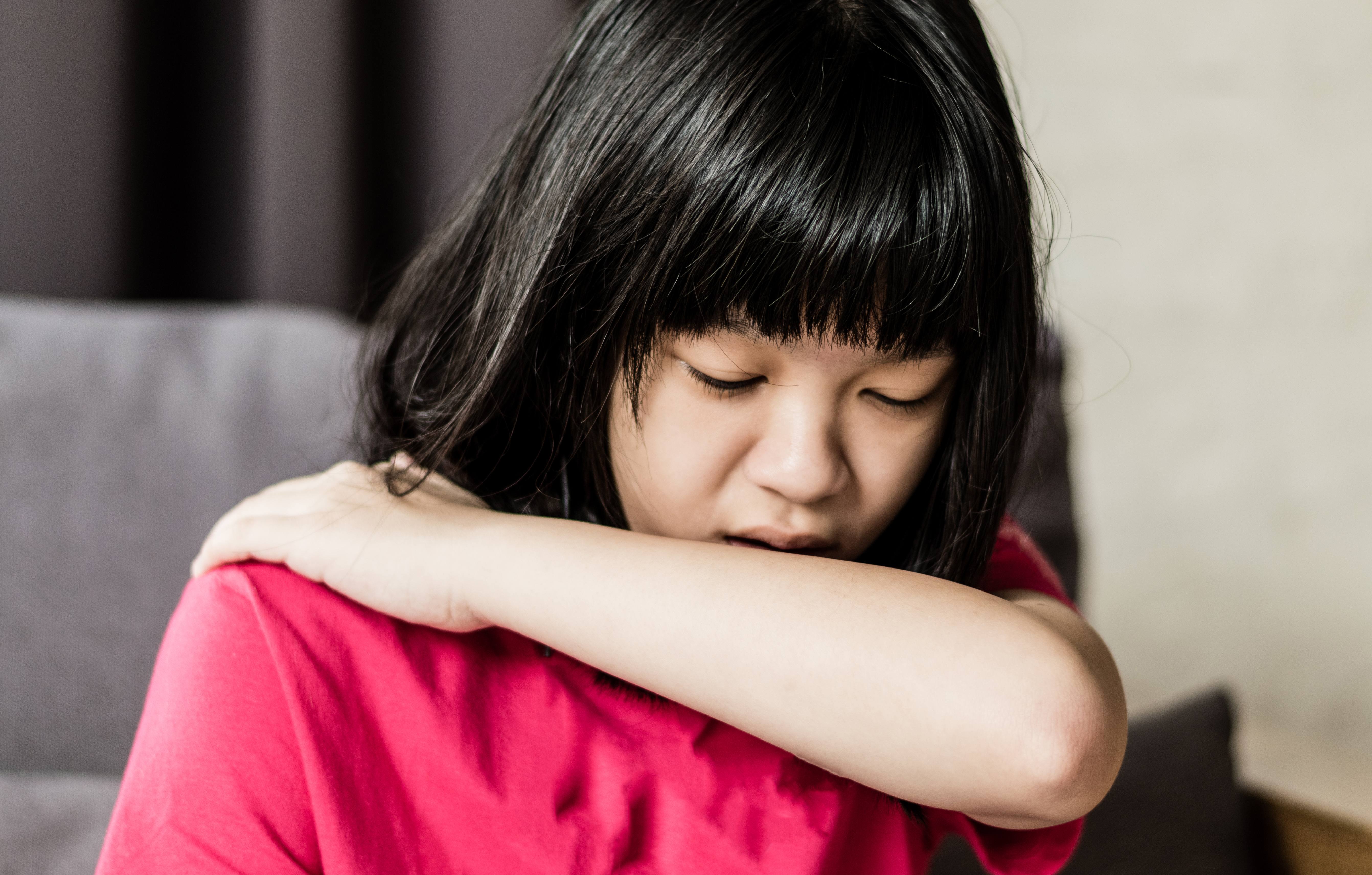 a young Asian girl who might be sick with a cold or the flu, is coughing into her elbow