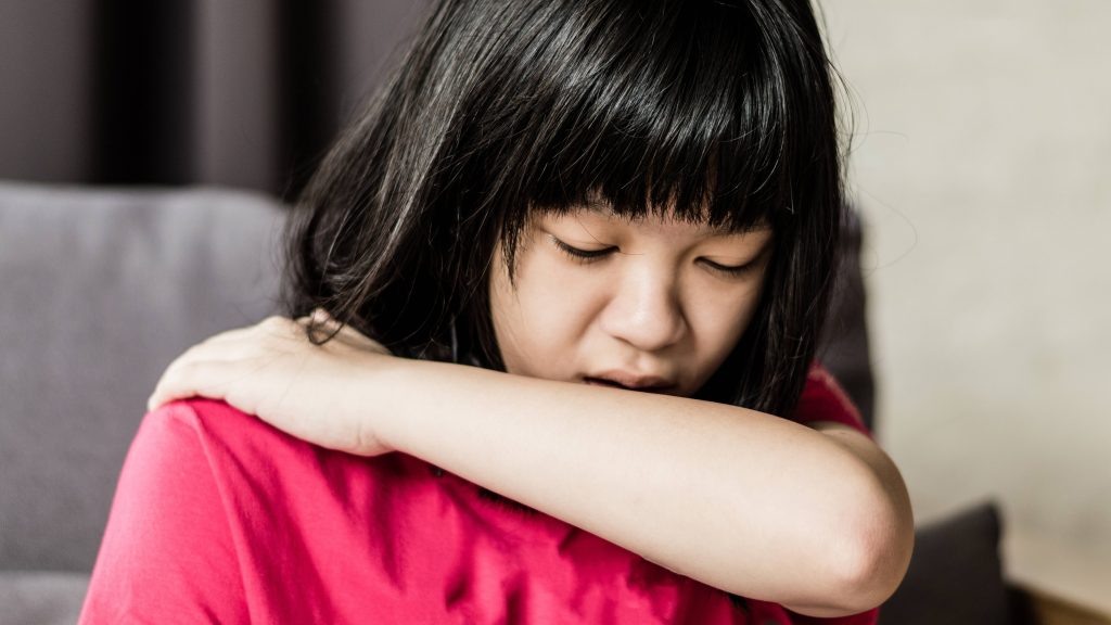 a young Asian girl who might be sick with a cold or the flu, is coughing into her elbow, respiratory illness