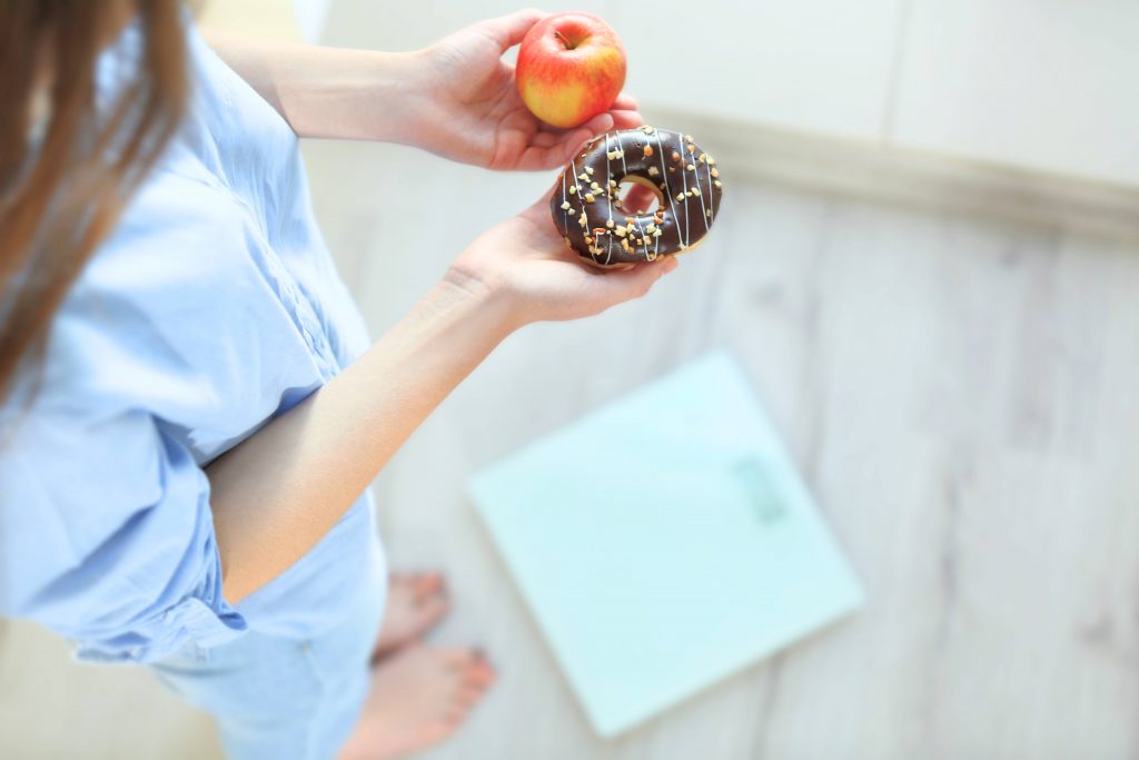a woman holding an apple in one hand a sprinkled donut in the other, while standing near a scale to weigh herself