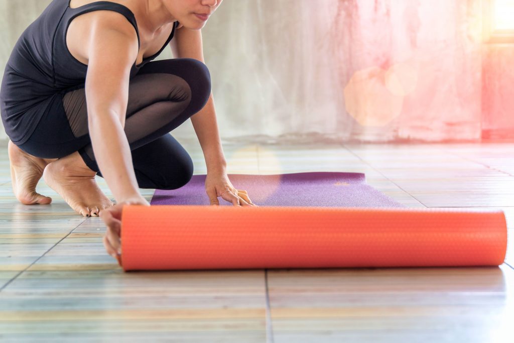 a woman in a yoga studio rolling out an exercise yoga mat on the floor with warm sunshine streaming in a window
