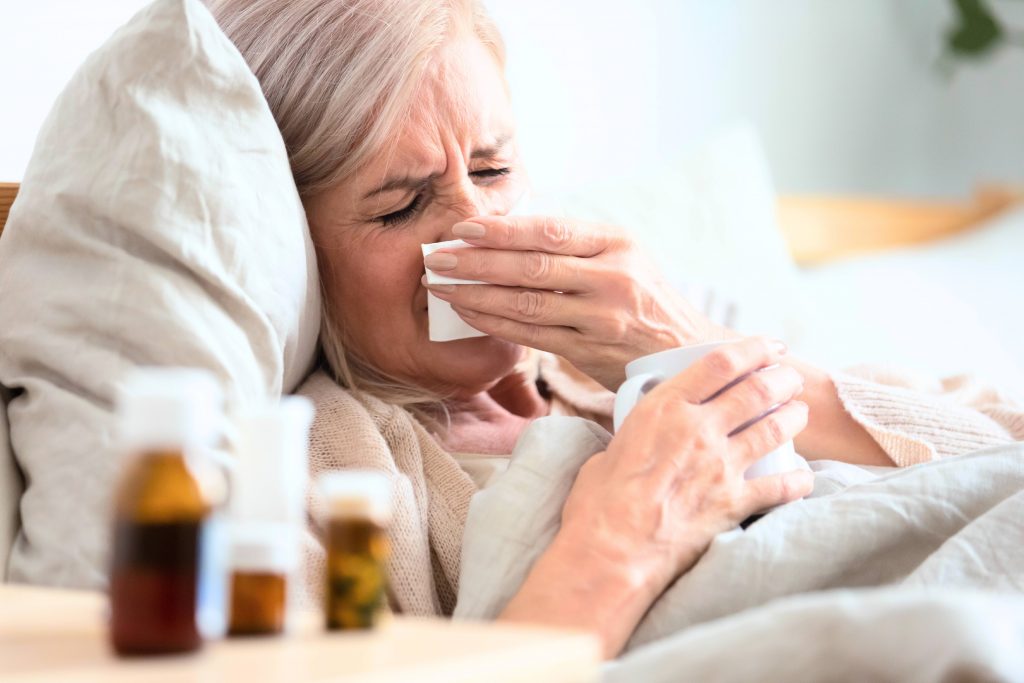 an older or middle-aged white woman sick in bed, with a cold or flu, holding a tissue sneezing and coughing