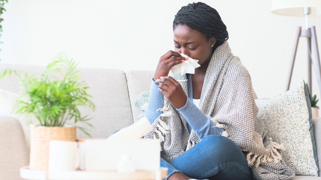 a young Black woman sitting on a couch wrapped in a blanket, blowing her nose and checking her temperature because she's sick with a cold or flu