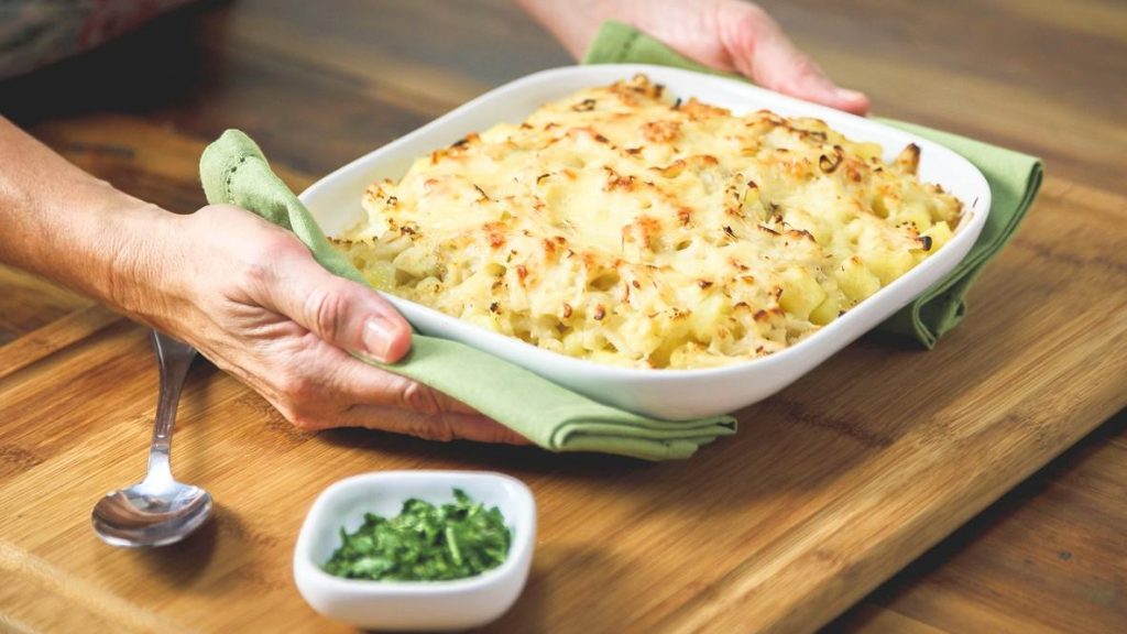 a person serving potato cauliflower au gratin in a white dish and holding the casserole dish with green napkins