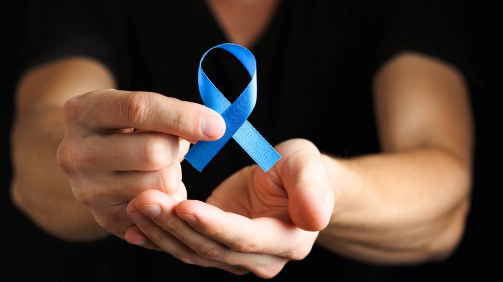 a white man in a dark shirt with a dark background holding a blue ribbon with his finger tips, representing colon cancer awareness