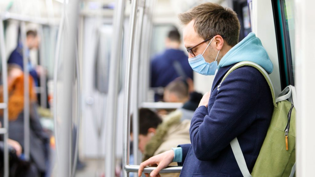 a young white man wearing glasses and carrying a backpack on a subway train, looking sick or ill and wearing a face mask to limit transfer of germs and infectious diseases