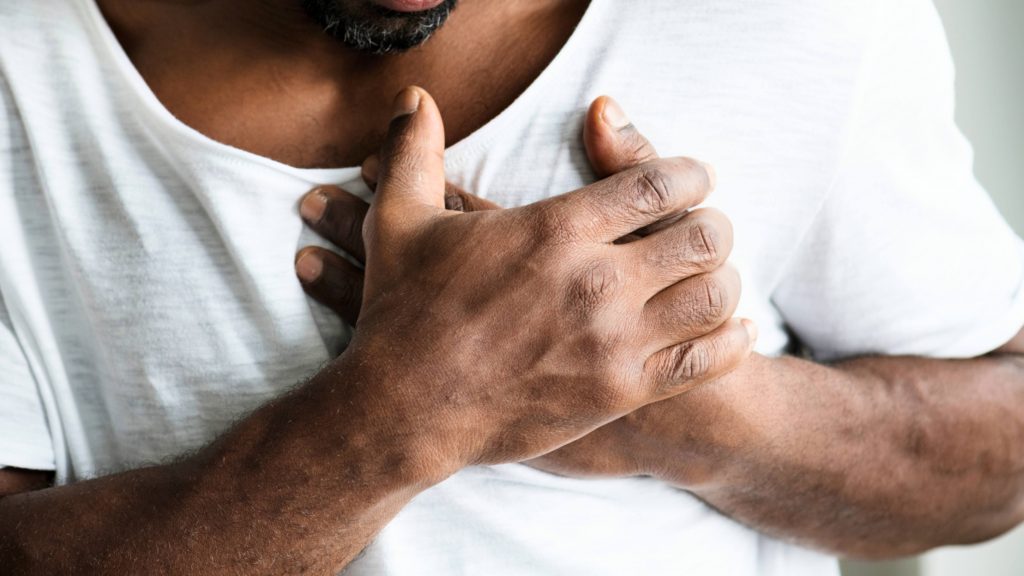 a closeup of a Black man in a t-shirt grasping his chest as if he's having heart pain or a heart attack