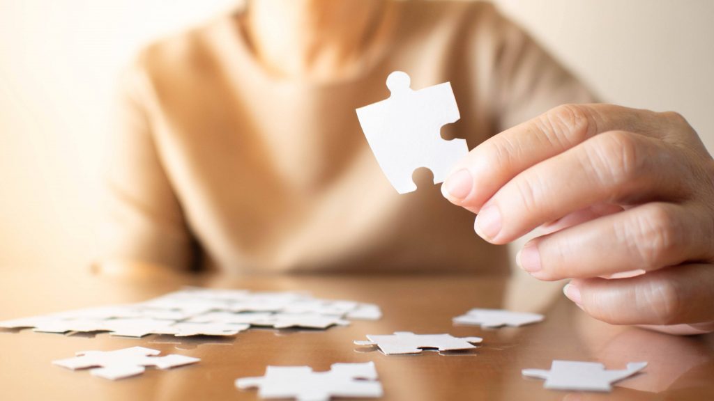 Elderly female hand trying to connect pieces of white jigsaw puzzle on wooden table. 