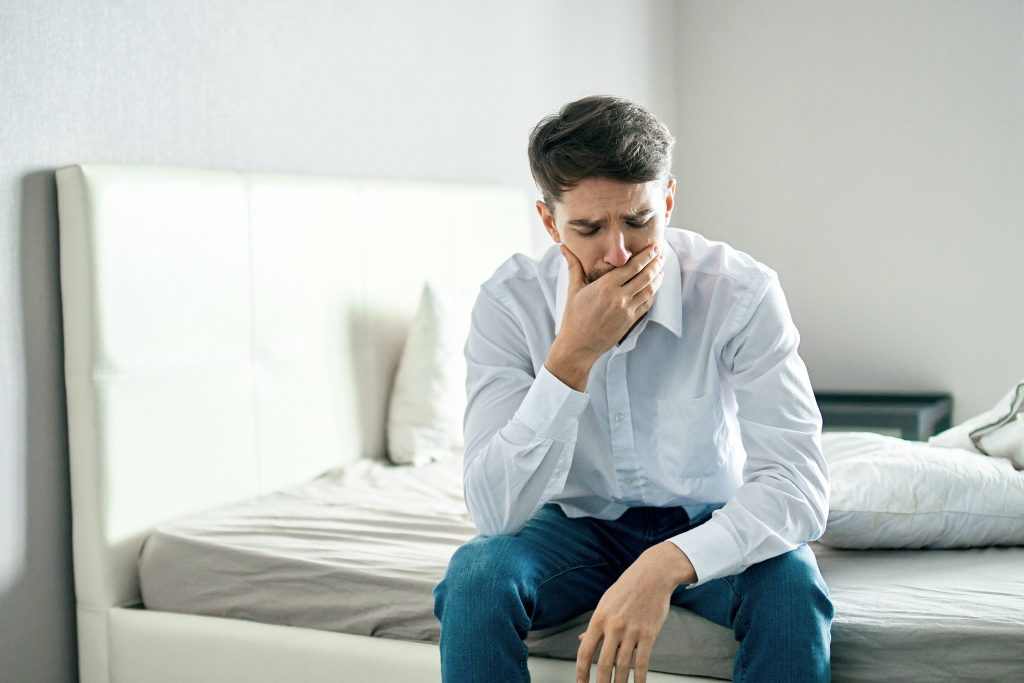 a young white man sitting on the edge of a bed, with his hand on his face looking sad, concerned, worried, stressed and depressed
