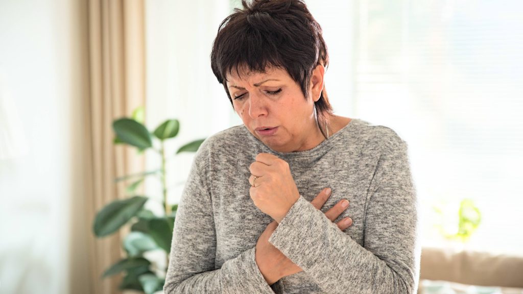 an older white woman, coughing, grimacing and looking unwell