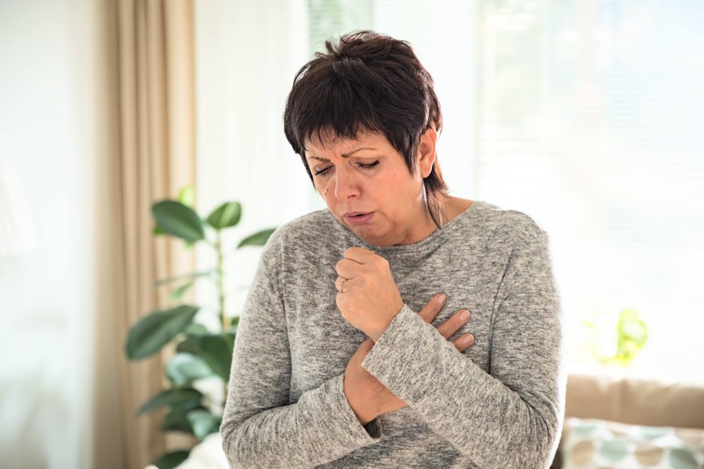 an older white woman, coughing, grimacing and looking unwell