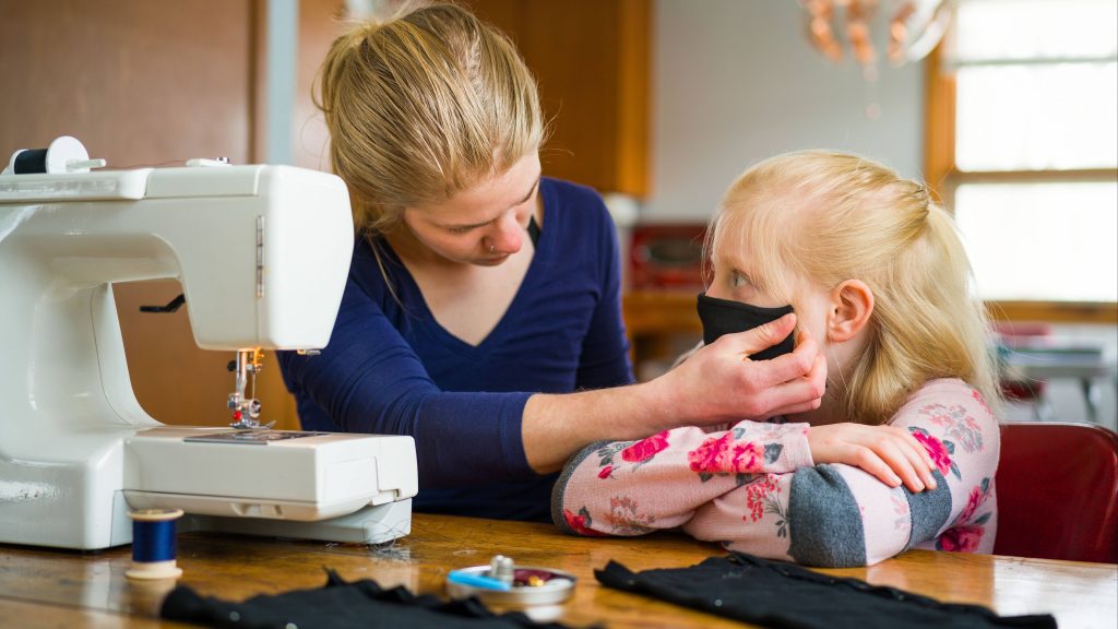 a young Caucasian woman at a sewing machine making face masks and with a little Caucasian girl sitting next to her and trying on one of the masks
