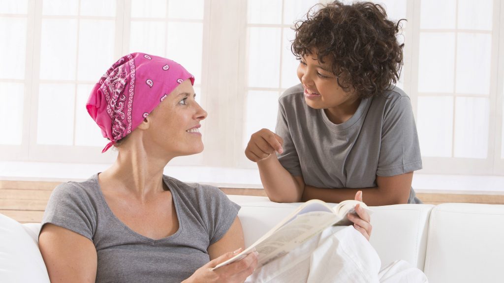 a Caucasian woman sitting on a couch, wearing a headscarf, perhaps a cancer patient having had chemotherapy, smiling and talking to a young African American or Latino boy, maybe her son