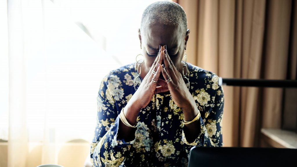 an African American woman sitting at a desk near a window with the light coming in and resting her head on her hands in a prayer-like position, perhaps worried or stressed