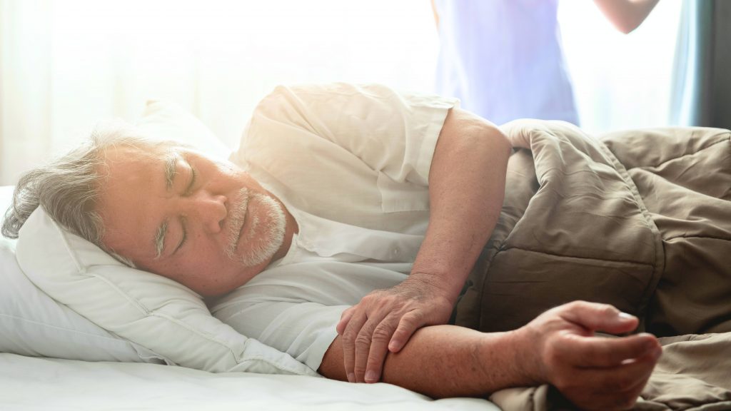 an elderly Asian or Latino man with a beard resting, sleeping under a blanket in bed, perhaps sick with a caregiver in the background opening a curtain
