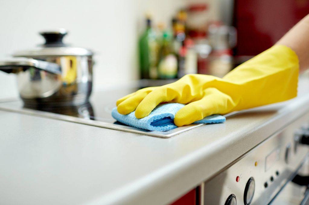 a person wearing a yellow rubber cleaning glove and wiping a stove top with a blue sponge