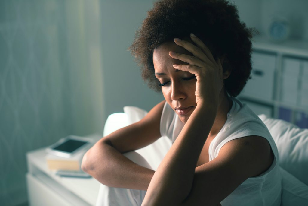 a young African American woman trying to sleep and sitting in bed in a dark room, with her eyes closed holding her hand on her forehead, perhaps suffering from insomnia, sadness, depression, worry. anxiety, stress