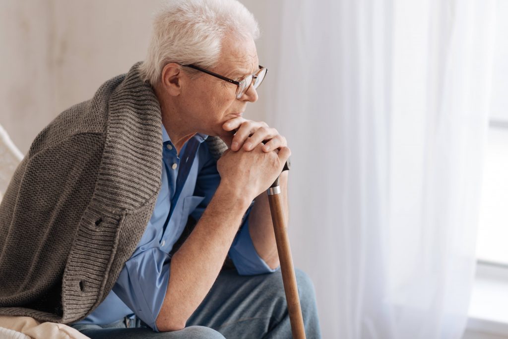 an older Caucasian man with glasses, leaning on a cane while sitting in a chair near a window and looking concerned, worried, thoughtful
