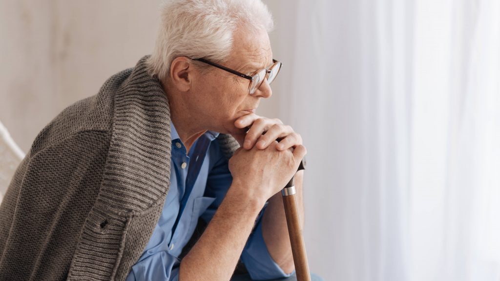 an older Caucasian man with glasses, leaning on a cane while sitting in a chair near a window and looking concerned, worried, thoughtful