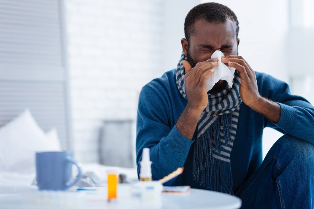 An African American man in blue jeans and a blue sweater blowing his nose into a tissue or sneezing, feeling sick with a cold, allergies or a flu bug