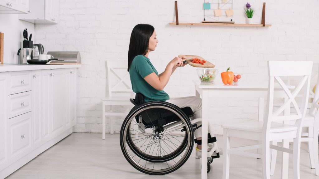 Vista lateral de una mujer discapacitada que está sentada en una silla de ruedas y cerca de una mesa preparando una ensalada