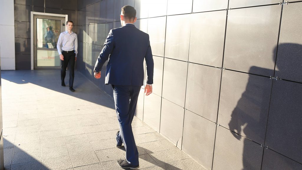 two Caucasian men in business clothes outside in the sunshine walking toward each other, keeping a distance between them