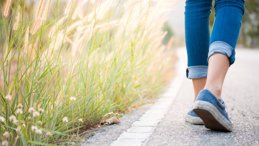 a closeup from behind of a Caucasian woman's leg and feet, wearing jeans and tennis shoes walking down a paved road beside tall grass