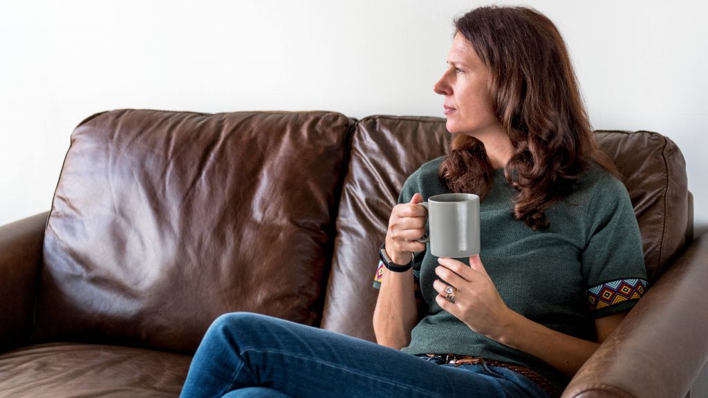 a middle-aged Caucasian woman in jeans sitting on a brown couch, holding a coffee mug, looking thoughtfully and seriously out a window, seeming calm