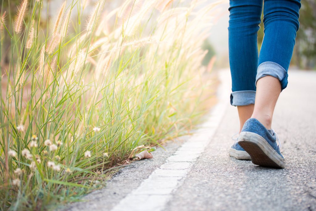 a closeup from behind of a Caucasian woman's leg and feet, wearing jeans and tennis shoes walking down a paved road beside tall grass