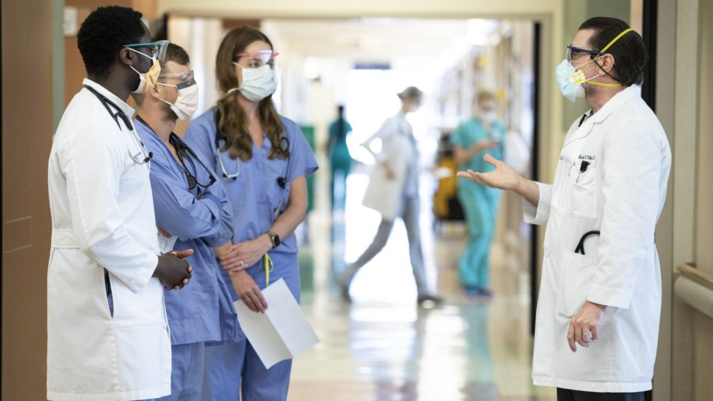 Mayo Clinic medical personel in scrubs, white jackets and protective face masks in a hospital corridor having a conversation