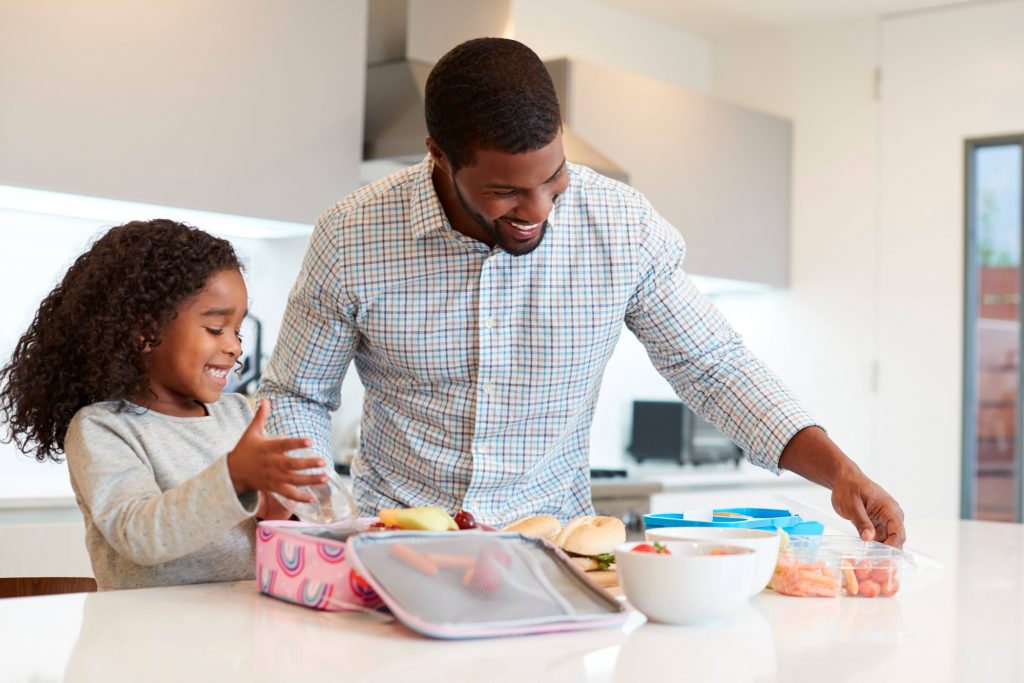 a Black man in a kitchen helping a little Black girl, perhaps his daughter, packing healthy food into her lunch bag - both of them smiling and happy