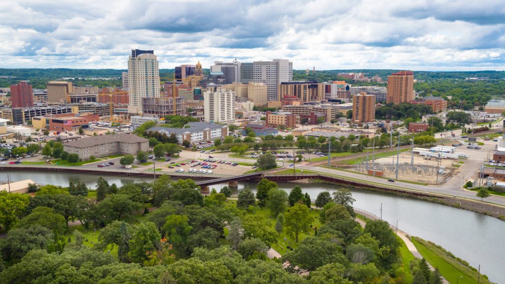 a landscape photo of downtown Rochester Minnesota, including the Mayo Clinic campus with a blue sky, some clouds and the Zumbro River with green trees in the foreground