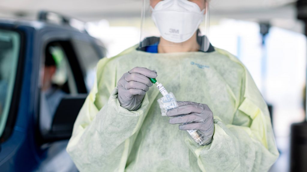 a medical staff person in PPE, mask, shield, gloves, gown, holding a COVID-19 test in her hands, after testing a patient in a nearby car