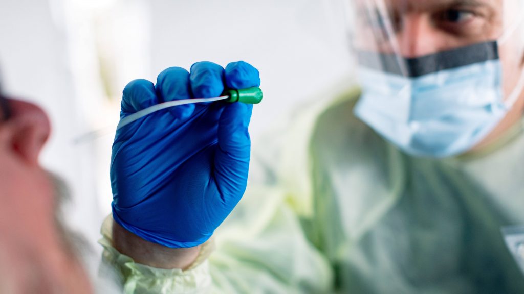 a patient being swabbed and tested for COVID-19 through a care window by a health care staff person wearing a face mask, blue gloves, face shield and protective gown