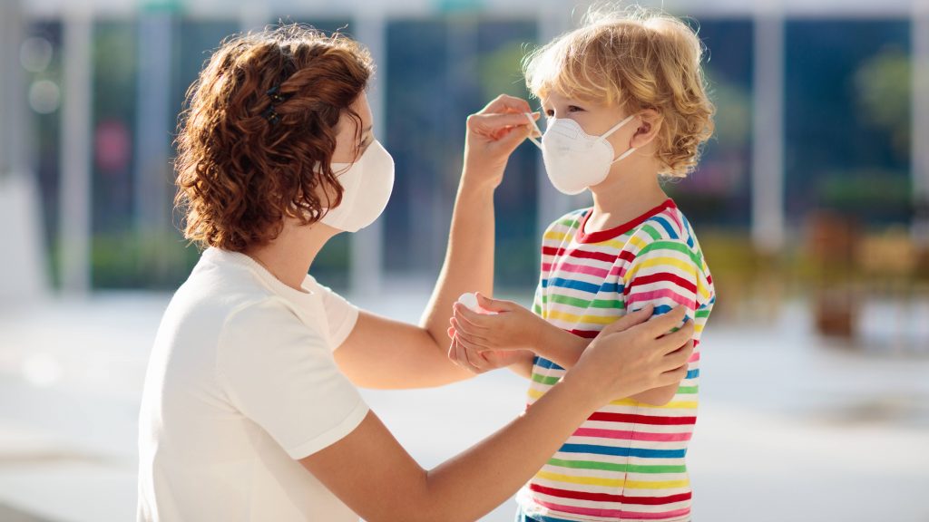 a white adult woman, perhaps a mother, wearing a mask and adjust the face mask on a young child