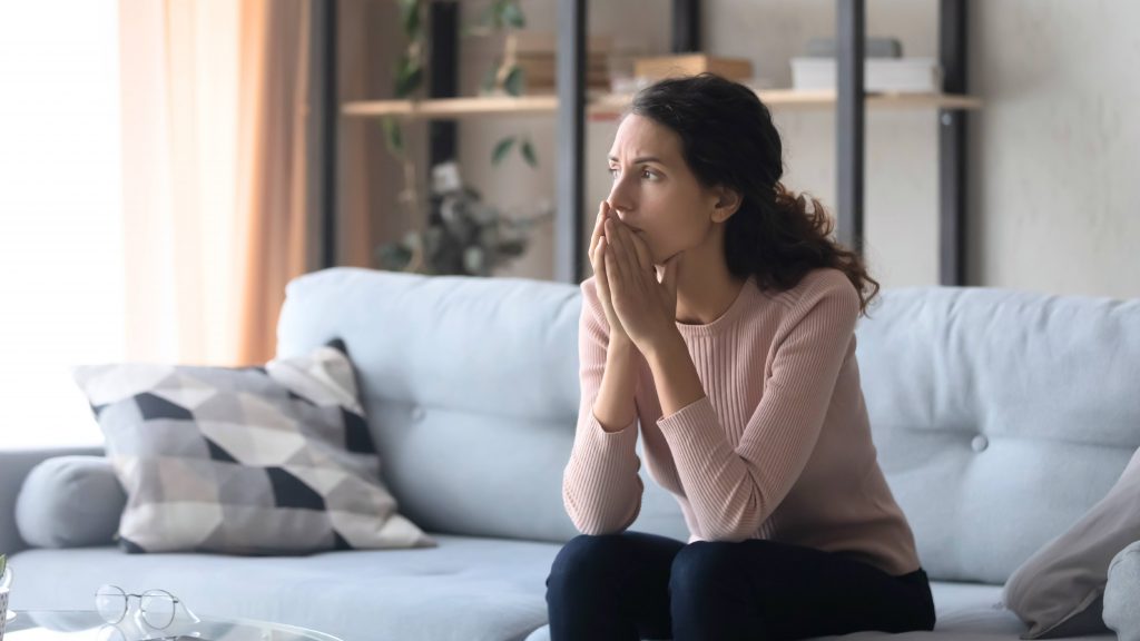 a white woman sitting on a couch looking out a window, crossing her hands and looking sad, worried, concerned
