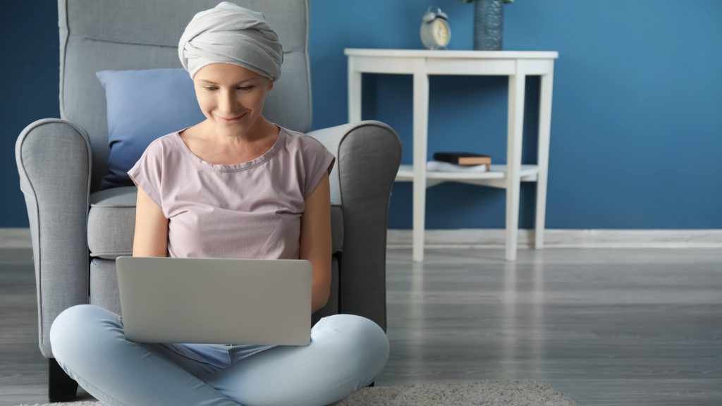 a young white woman wearing a head scarf and sitting on the floor in her home, smiling and working on a computer laptop