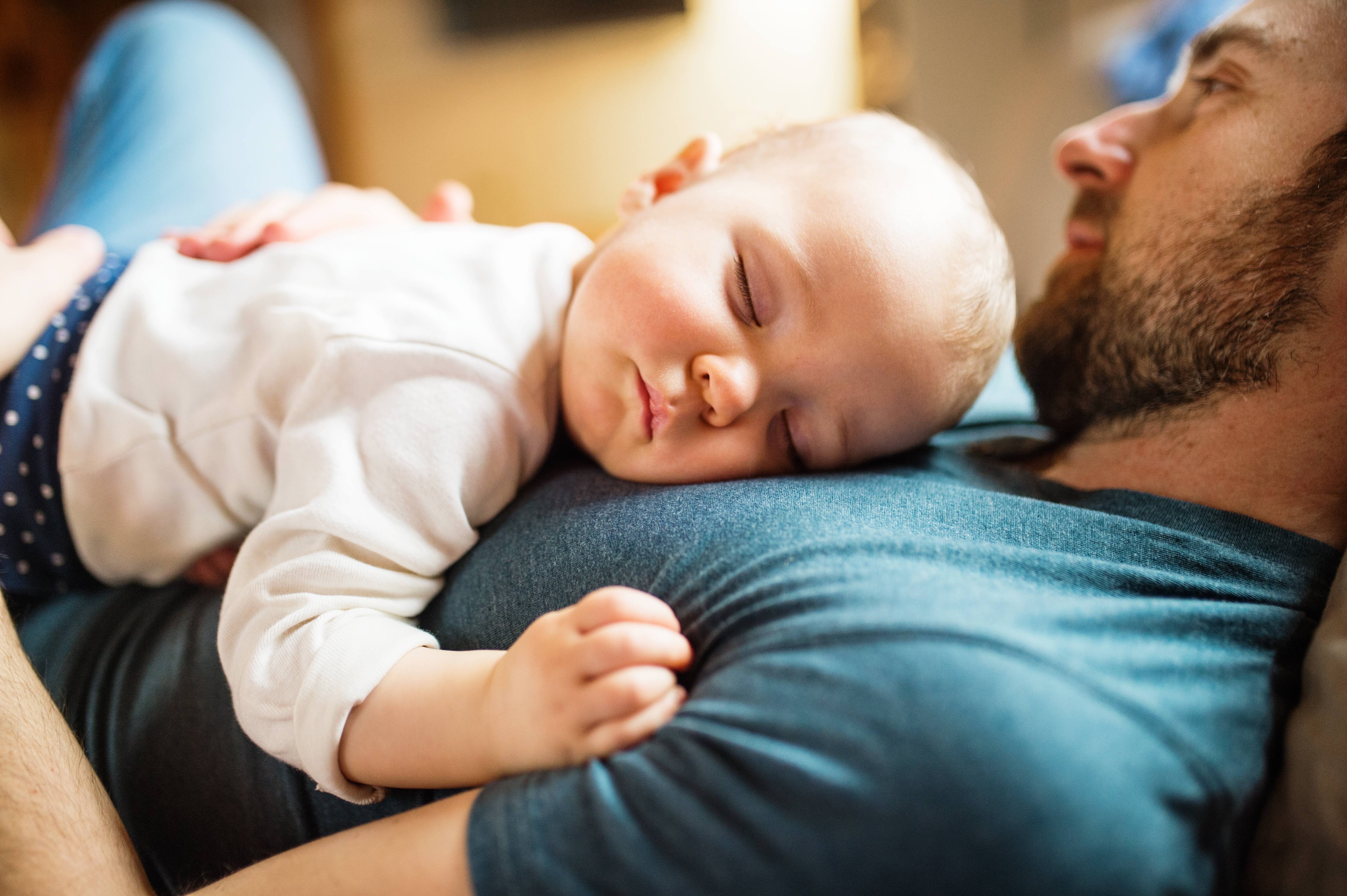a white man in a blue t-shirt resing on his back with an white infant, baby sleeping on the man's chest
