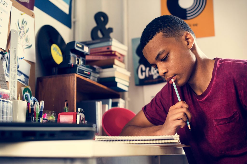 a young Black teenage boy in a t-shirt working at a desk, with a notebook and a pencil, perhaps doing homework in his bedroom or college dorm room