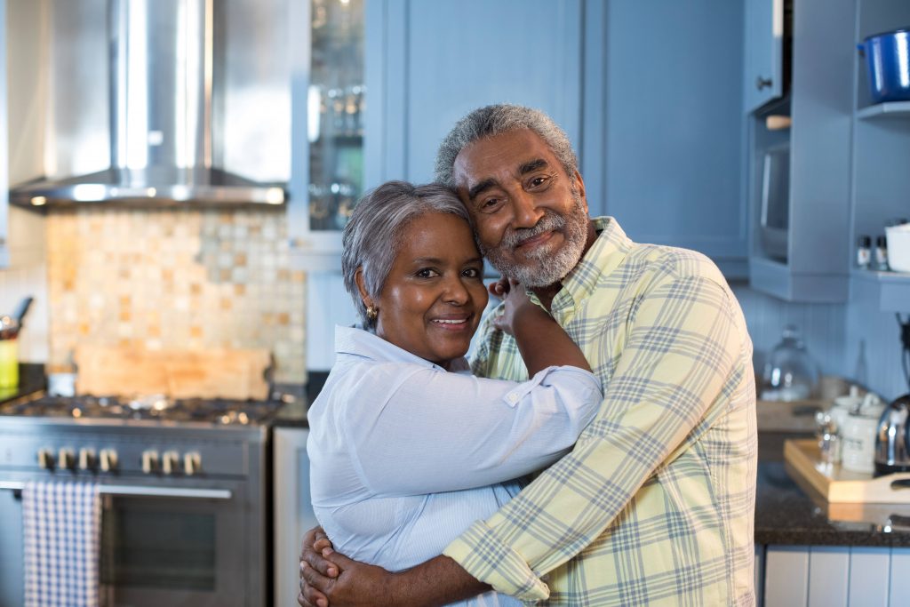 a middle-aged couple, perhaps Black or Latino with little brown skin tones, smiling, happy and hugging in a kitchen