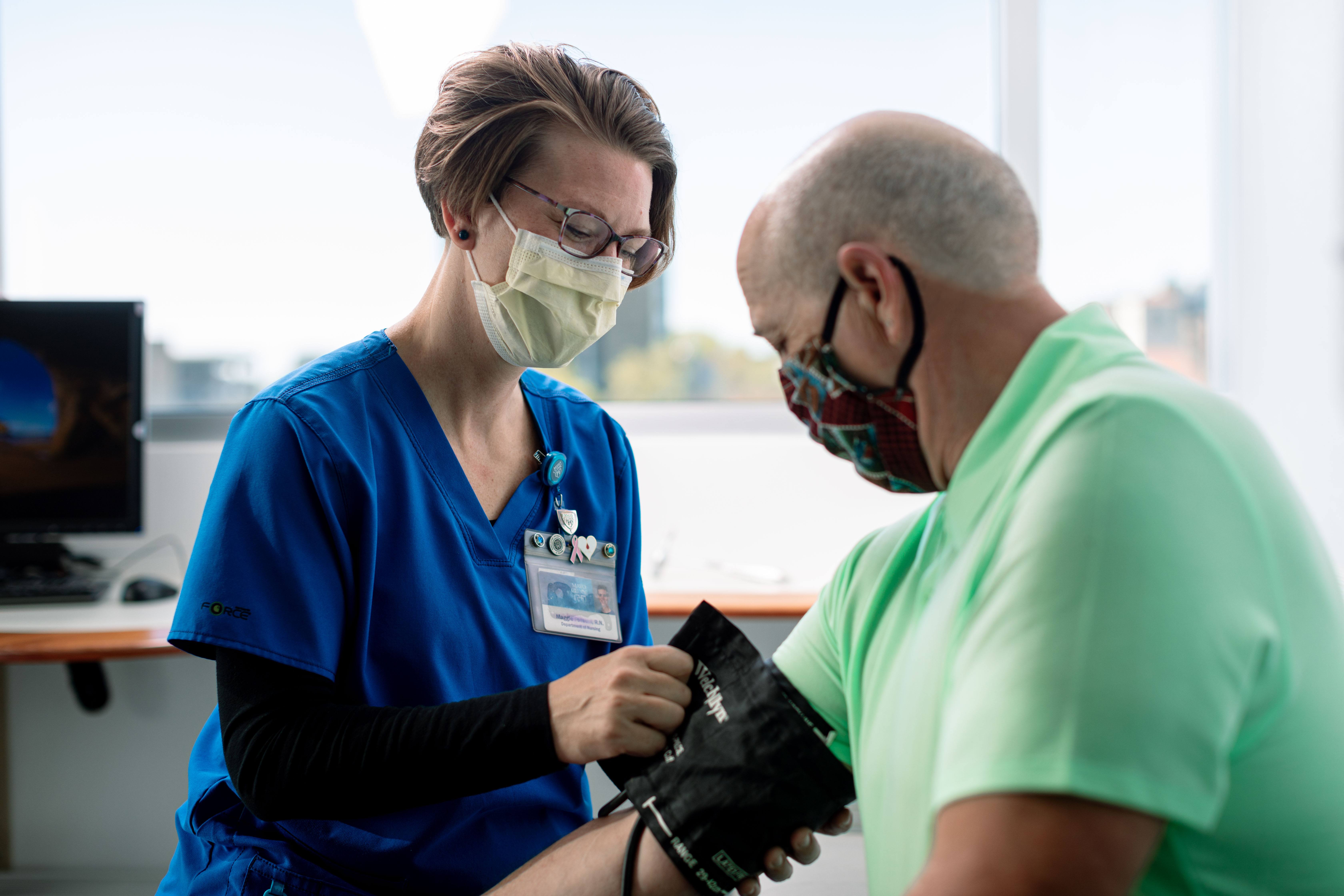 a white woman, Mayo Clinic health care worker in blue scrubs and wearing a mask is taking a white man's blood pressure, who is also wearing a mask