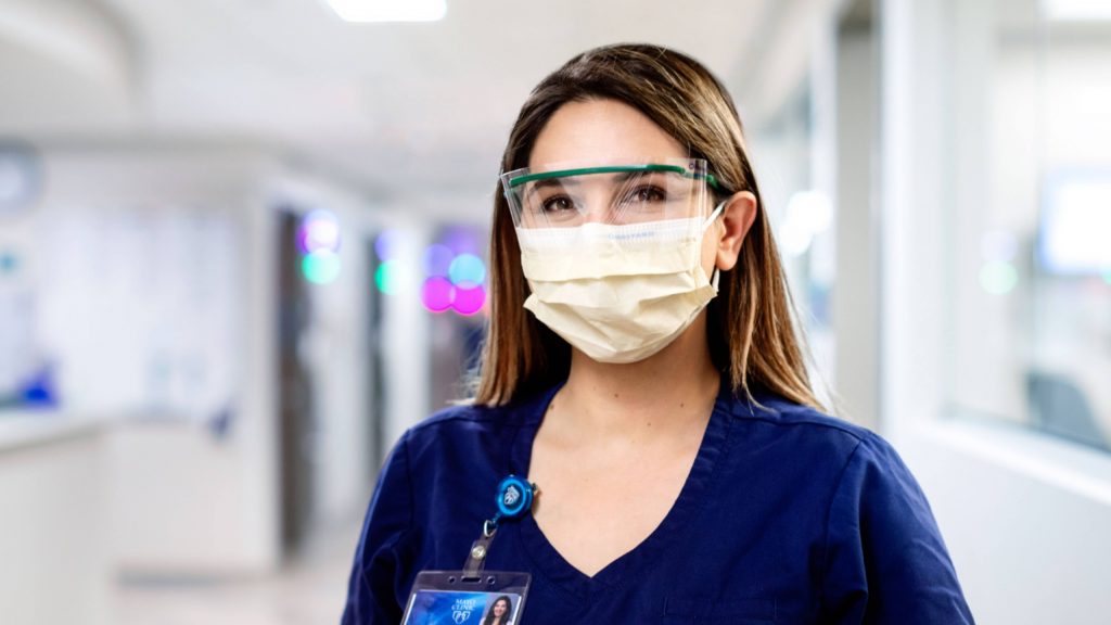 a white or Latino woman in Mayo Clinic medical blue scrubs and wearing eye protection and a face mask in the hospital corridor