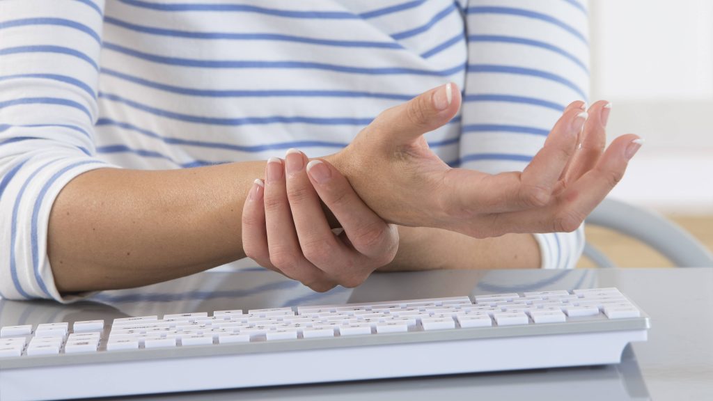 a white woman in a striped shirt near a computer keyboard holding her wrist because it's injured, sore, in pain