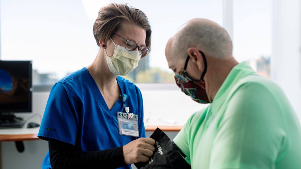 a white woman, Mayo Clinic health care worker in blue scrubs and wearing a mask is taking a white man's blood pressure, who is also wearing a mask