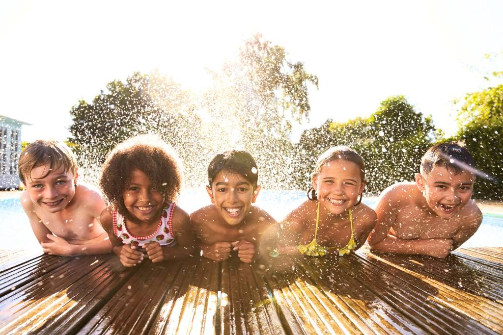 five smiling tweenage girls and boys leaning on the edge of a swimming pool with water splashing behind them