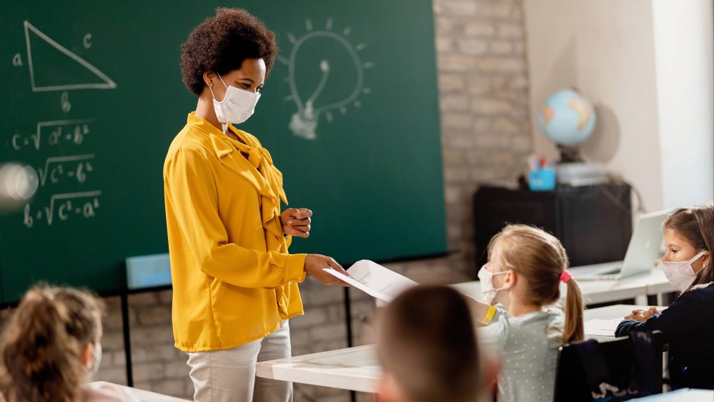 a Black woman teacher wearing a mask and standing in front of a classroom of students wearing face masks, with a chalkboard in the background