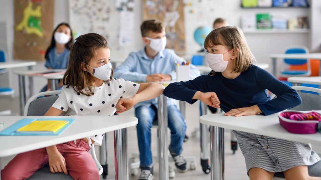 a classroom of elementary or middle-school aged children wearing masks and praticing social distancing, touching elbows