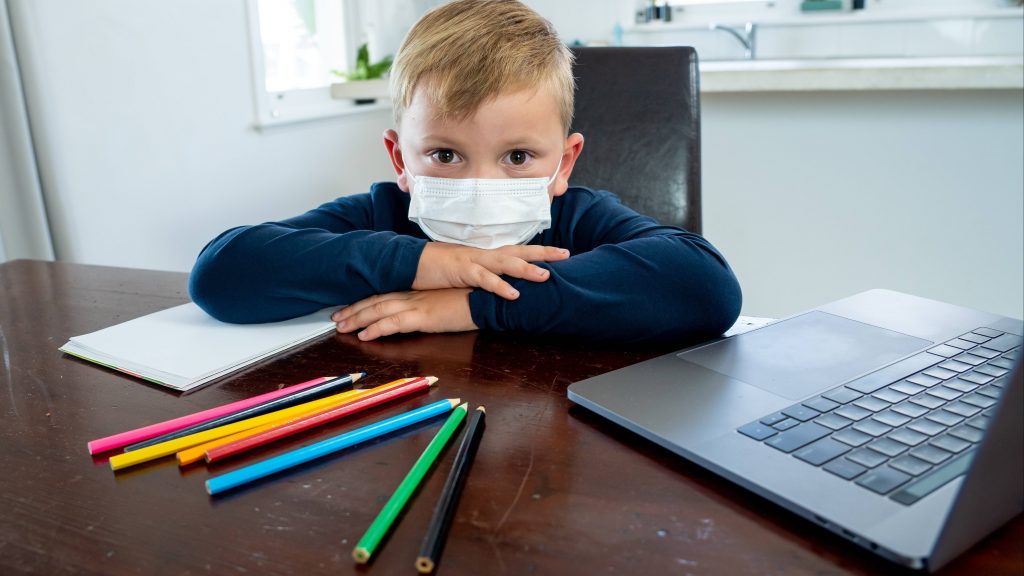a young white school aged boy looking sad and lonely, wearing a mask and sitting at a desk with paper, colored pencils and a laptop computer